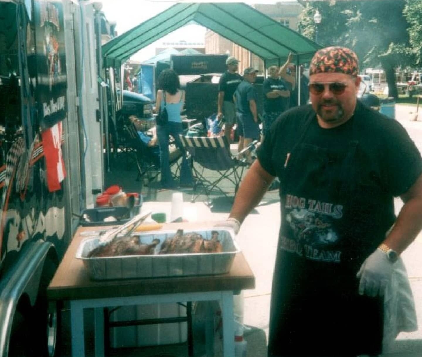 Chris Corrigan stands under a canopy at a barbecue competition wearing a Hog Tails BBQ Team apron, sunglasses, and a bandana, tending ribs beside a tray of cooked meat while other teams work in the background.